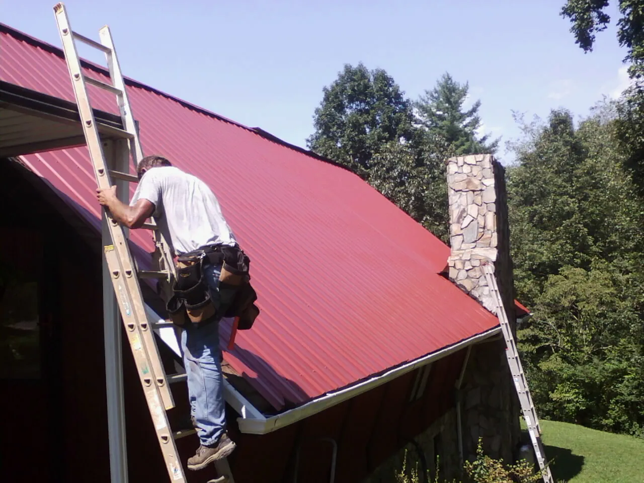 Roofer on a ladder beside a red metal roof and stone chimney — Soffit & Fascia Repair by Roofing Brothers in Cambridge